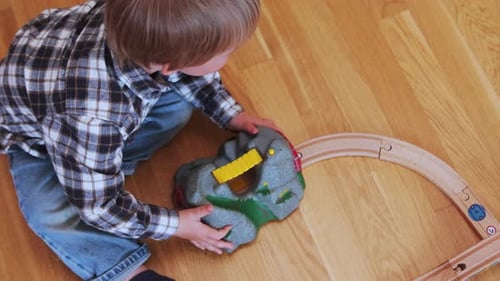 Toddler adding a tunnel to a wooden train set while sitting on the floor. Shot from above