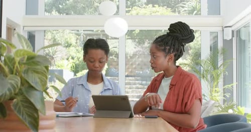 Une mère afro-américaine et une fille adolescente à table pointant vers une tablette, sa fille prenant des notes