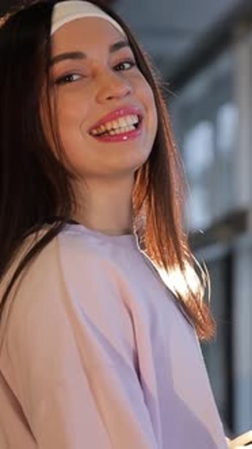A Young Woman with Long Layered Hair and a Headband Stands By a Gym Window Vertical Video