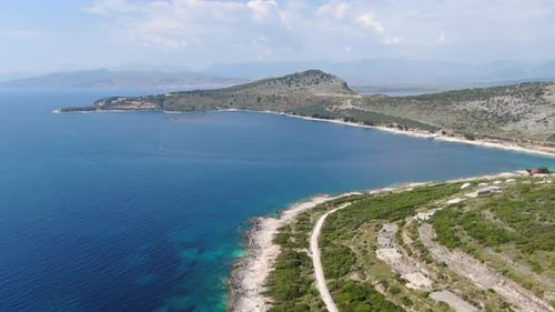 Drohnenblick in Albanien beim Flug über blaues Wasser, Meer neben grüner Berglandschaft, sonniger Tag in Ksa