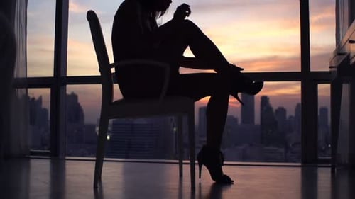 Silhouette of woman putting on heels in city apartment