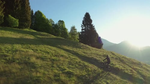 Drone shot of a young athletic man on MTB e-bike pedalling up the hill with green grass at sunset.