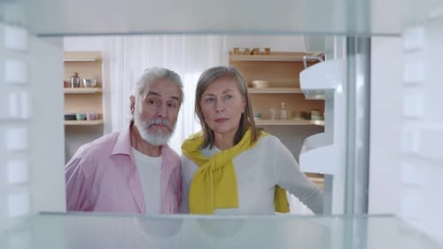 A Couple Peers Into an Empty Fridge Expressing Curiosity and Disappointment