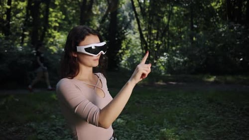 In a Blurred Nature Background an Outdoors Game is Being Played By a Young Woman in VR Glasses