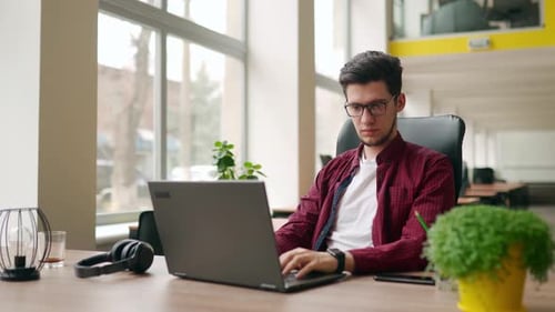 Junger stilvoller Freelancer, der mit dem Laptop arbeitet und dann die Kamera betrachtet. Büroangestellter, der auf dem PC tippt