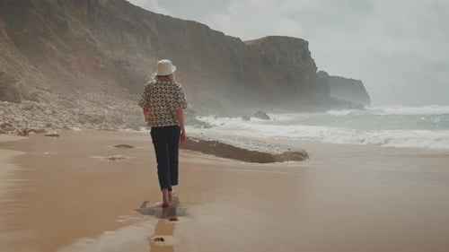 Back view of tourist girl walking barefoot on the sandy beach by the blue ocean.