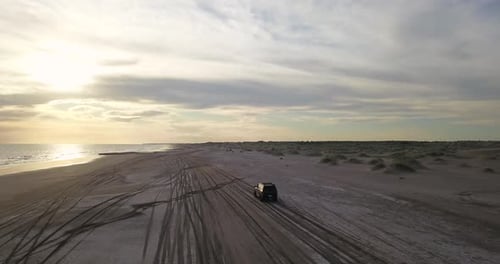 Car Driving On The Beach Along The Sea And Sand Dunes At Sunset. - aerial shot