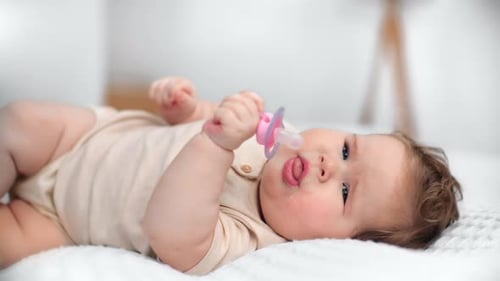 Säugling trägt einen beigefarbenen Body, ruht auf einer weißen Decke, schnappt sich einen Schnuller und macht sich verspielt
