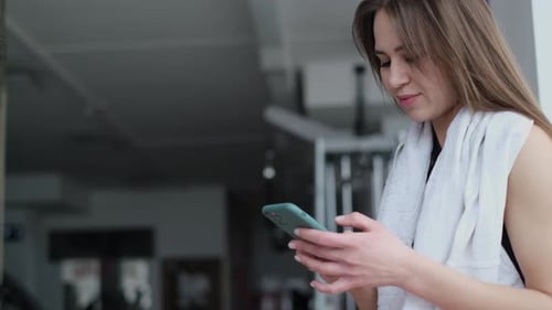 The Girl After Training in the Gym She is Tired with a Towel Around Her Neck Uses a Smartphone