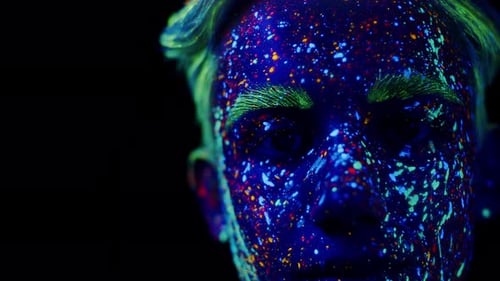 Face of a boy covered in neon paint glowing in ultraviolet light. Young man in fluorescent makeup lo