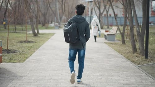 Man in Casual Clothing with Backpack Walking Alone Along Paved Park Path on Overcast Day