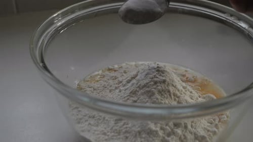 Male hand sprinkling baking powder from metal spoon into mix of flour and egg in glass bowl. Slow mo