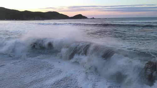 Vista de drone da onda de tempestade caindo na praia arenosa e nas rochas ao nascer do sol