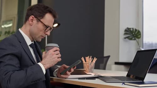 Male Employee Drinking Coffee and Using Smartphone at Office