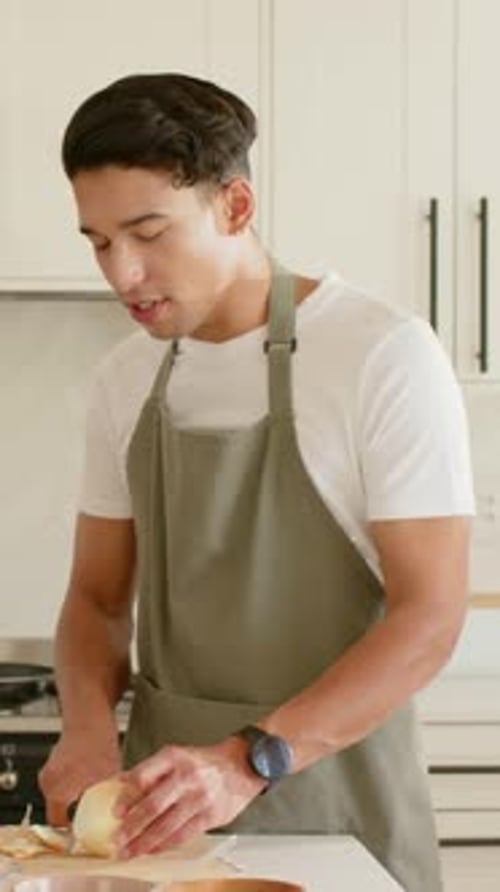 Vertical video: Starting cook in olive apron peeling onion on board for meal at home, showing onion