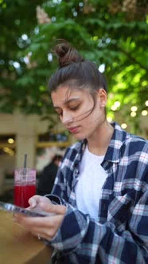 With a Café Terrace Setting a Pretty Girl Holds a Smartphone and Sips a Cocktail on a Summer Day