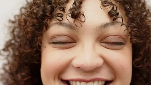 Opening closeup, woman beaming at camera on off-white background, closing, reopening eyes, smiling