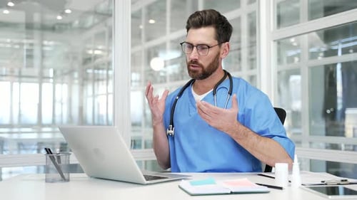 Confident doctor in blue coat talking on video call using laptop sitting in hospital office.