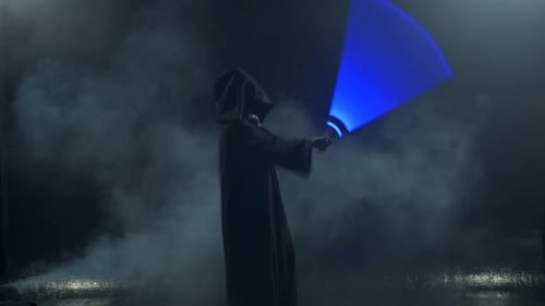 Young Man in Jedi Cosplay Costume with Lightsaber Battle on Black Background in Smoke and Rain Slow