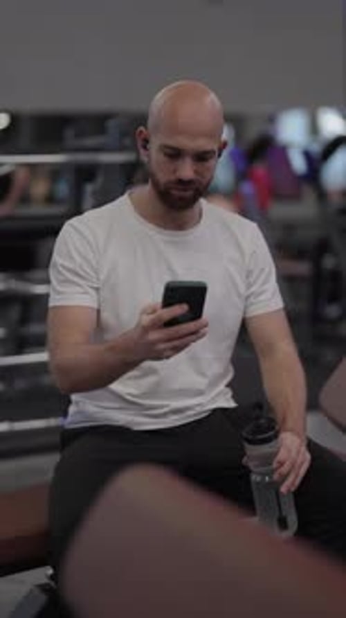 Bald Man Using Smartphone and Drinking Water After Workout at the Gym