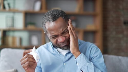 Sick mature african american male suffering from cold and headache sitting on sofa in the living