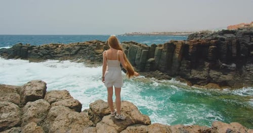 Rear view of young slim blonde woman, white dress, very long hair, standing on rocky ocean beach, sl