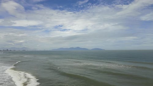 Waves in the ocean breaking towards the coast with big fluffy clouds in the sky and mountains in the