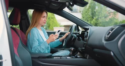 Beautiful young woman driver in smart clothes using mobile phone while sitting in luxury car. Young