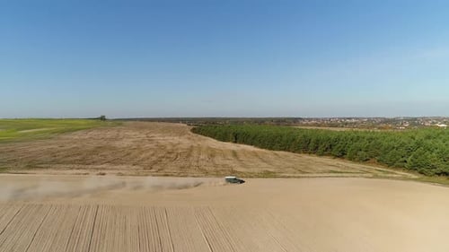 A Green SUV Drives Quickly Along a Dirt Road Between Two Fields and a Forest