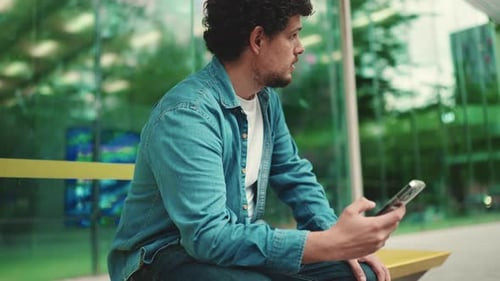 Close-up, man sits at bus stop waiting for bus and uses mobile phone