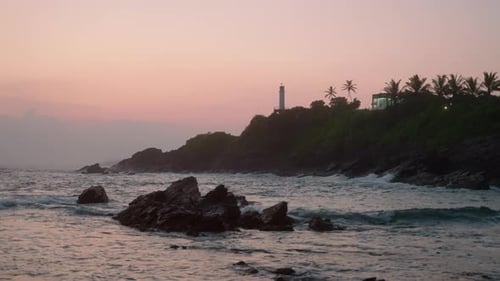 Silhouetted Lighthouse Stands on Hill at Dusk Waves Crash on Rocky Shore Beneath Cotton Candy Sky