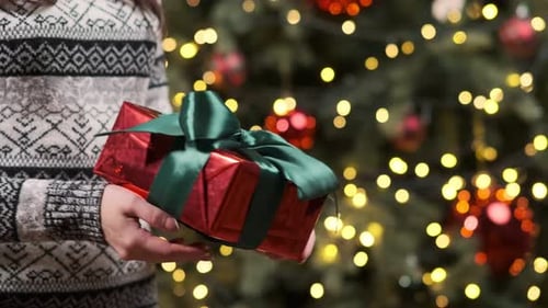 Nahaufnahme eines Mädchens in einem Weihnachtspullover, das eine rote Geschenkbox mit einem grünen Band in den Händen hält.
B