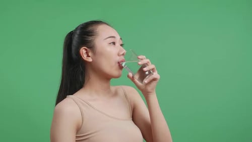 Seitenporträt einer asiatischen Frau, die Wasser trinkt, während sie auf grünem Hintergrund steht