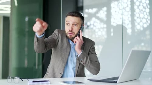 Angry dissatisfied businessman arguing talking on the phone sitting at workplace in business office.