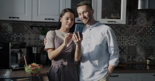 Loving Couple Smiles While Looking at Smartphone in Kitchen