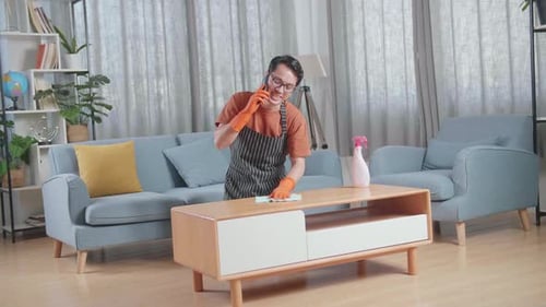 Asian Male Housekeeper With An Apron Talking On Smartphone While Cleaning The Table By The Spray