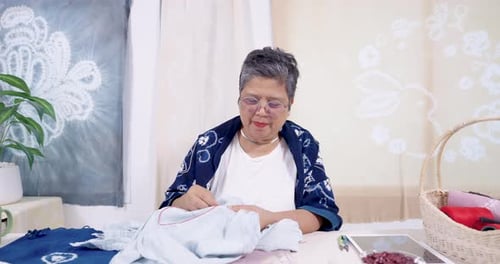 Ältere asiatische Frau Kunsthandwerkerin lächelt, zieht roten Faden durch Denim, zieht fest, widersteht Stich, hält