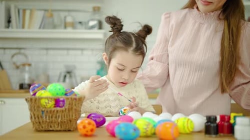 A happy mother and daughter enjoy painting colorful Easter eggs at a kitchen table together.