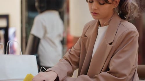 Woman Seated at Mall Table Flipping Catalog Pages and Reviewing Product Images Shopping Bags Beside