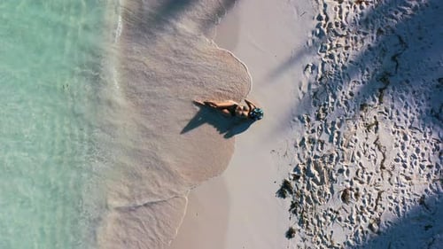 aerial top down of young woman in bikini and hat laying on tropical white sand beach shoreline with