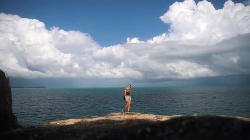 Young blond tourist walks over stones and enjoys the beautiful view of the vast sea in Thailand on a