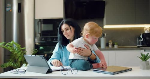 Woman Holding Toddler Working at Kitchen Table