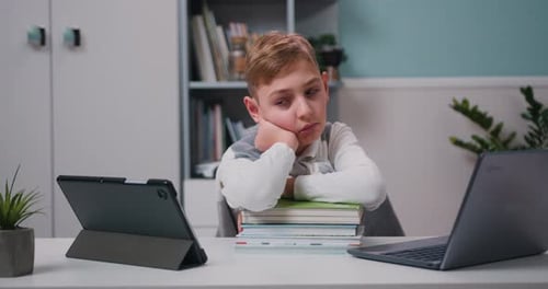Tired school boy with sad face sitting at the table with notebooks and laptop during online studying