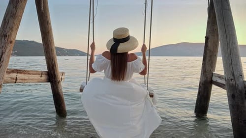 Slow-motion beauty of an ocean beach, where a young woman in a dress sways on a swing