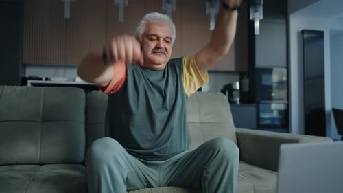 Retrato de hombre mayor sano entrenando mediante videotutoriales en línea levantando mancuernas sentado en el sofá