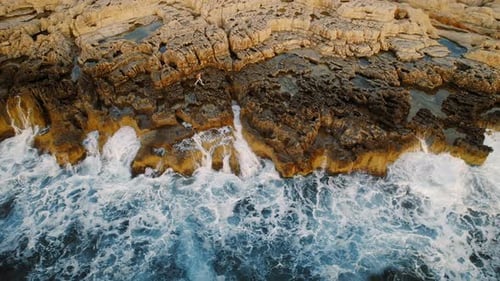 Person climbing rugged coastal rocks above crashing ocean waves at sunset