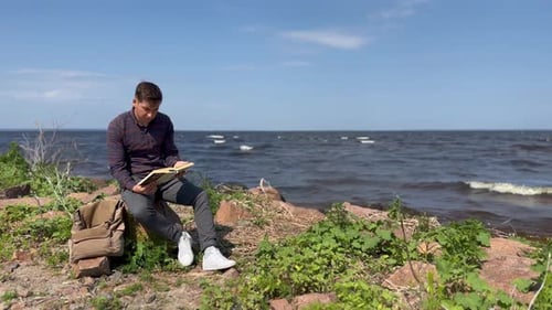 A man sits on a stone by the sea and reads a book, on a sunny day