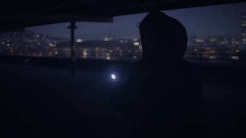 Hooded Figure with Flashlight in Dark City at Night