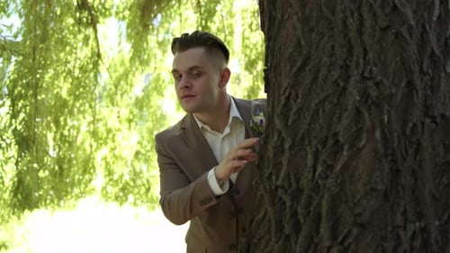 Bonito homem confiante de terno olha para trás de uma árvore e está feliz com o que viu, emoções