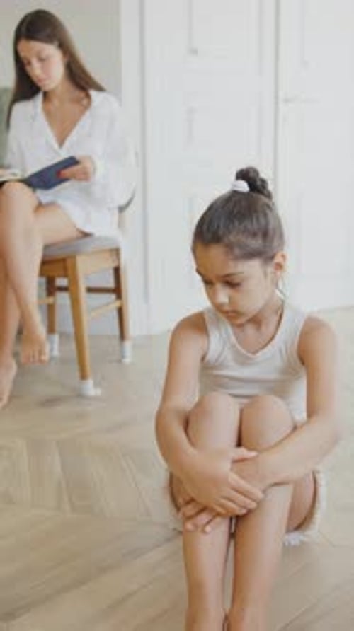 A Sad Little Girl Sits on the Floor with Her Knees Pulled Up and Hugging Them While a Therapist Sits